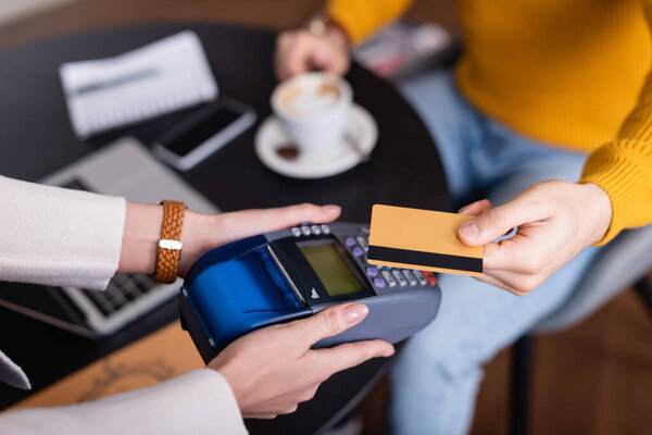 cropped view of waitress holding payment terminal near teleworker with credit card, blurred background