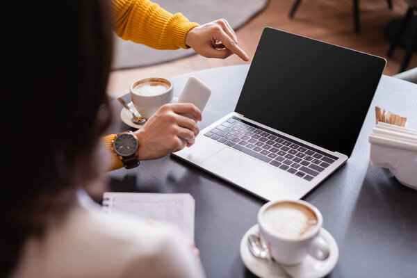 Человек со смартфоном указывает на ноутбук с пустым экраном рядом с кофе и друг на размытом переднем плане в ресторане 