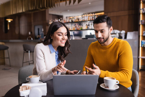 Мультиэтнические друзья смотрят на ноутбук возле кофе на размытом переднем плане в ресторане 