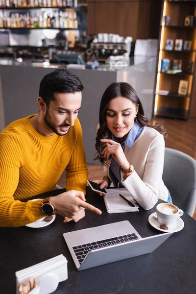 Arabian man pointing at laptop near friend with smartphone near coffee in restaurant