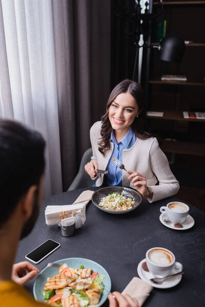 Smiling woman looking at boyfriend near dinner, coffee and smartphone on blurred foreground in restaurant 