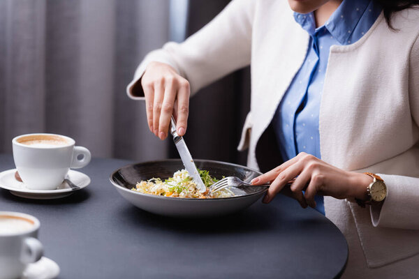 Cropped view of young woman cutting salad near cups of cappuccino on blurred foreground in restaurant 