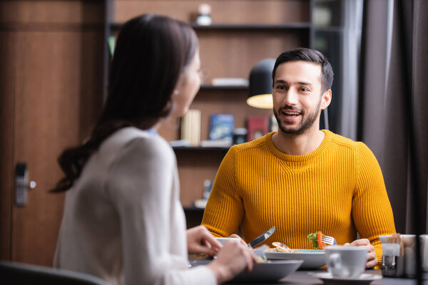 Smiling muslim man holding cutlery near tasty dinner with girlfriend on blurred foreground 