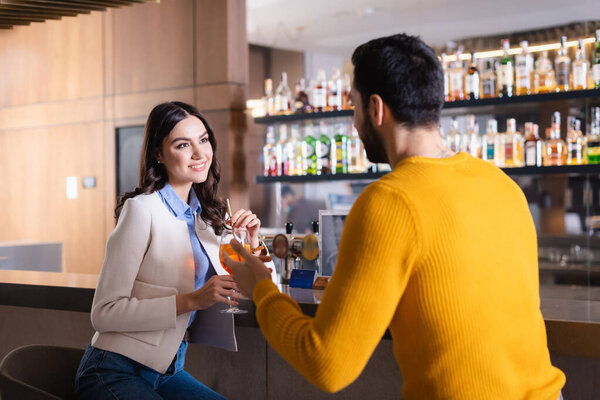 Smiling woman with cocktail looking at muslim friend on blurred foreground near bar counter in restaurant 