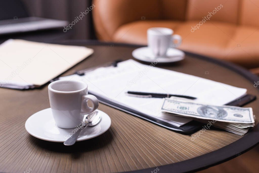 Cup of coffee near dollars and clipboard on blurred background on table 