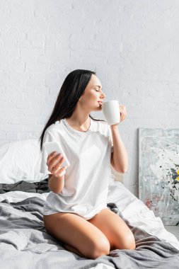 Fincanlı ve akıllı telefonlu genç bir kadın yatakta oturuyor. 