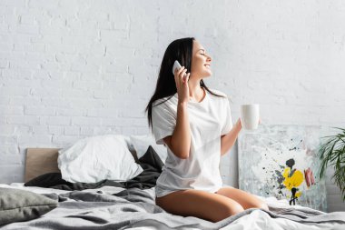 Bardaklı neşeli kadın yatakta akıllı telefonla konuşuyor. 