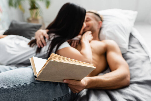 Book in hand of shirtless man kissing girlfriend on blurred background on bed 