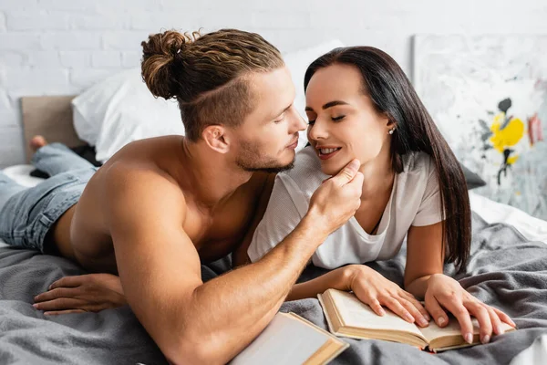 Shirtless Man Touching Chin Smiling Girlfriend Books Bed — Stock Photo, Image