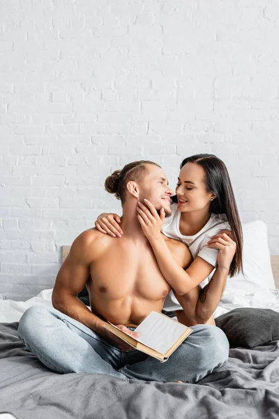 Sexy Man Holding Book Embracing Cheerful Girlfriend Bed — Stock Photo, Image