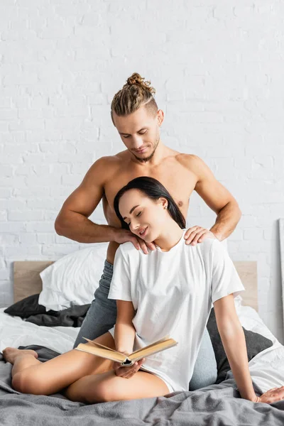 Muscular Man Touching Shoulders Smiling Girlfriend Book Bed — Stock Photo, Image