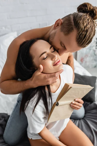 Hombre Sin Camisa Besando Novia Morena Con Libro Dormitorio — Foto de Stock