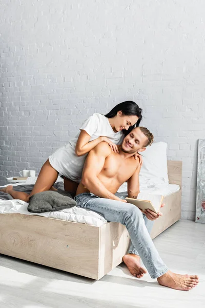 Mujer Morena Camiseta Abrazando Novio Sonriente Con Libro Cama — Foto de Stock