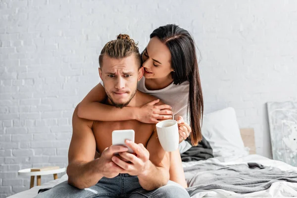 Mujer Sonriente Con Taza Abrazando Novio Confundido Usando Teléfono Inteligente — Foto de Stock