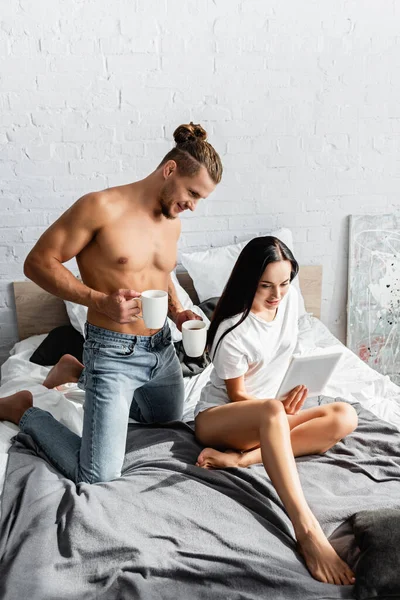 Sonriente Hombre Sosteniendo Tazas Cerca Novia Con Tableta Digital Cama — Foto de Stock