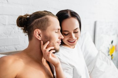 Gülümseyen kadın yatakta üstsüz adama dokunuyor. 