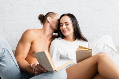 Seksi bir adam gülümseyen kız arkadaşını öpüyor. Önplanda bulanık bir kitapla. 