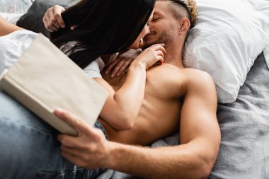 Yatakta kız arkadaşını öperken ön planda bulanık bir kitap tutan üstsüz bir adam. 