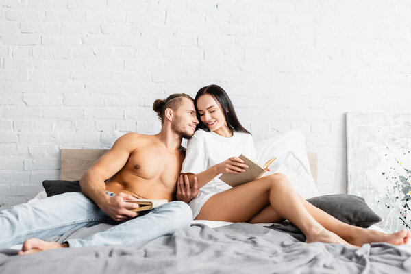 Smiling woman holding book near sexy man in jeans on bed 