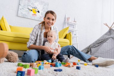 cheerful woman looking at camera while playing with colorful cubes on carpet with son clipart