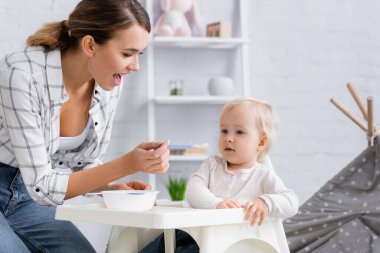 Açık ağızlı genç bir anne çocuk koltuğunda oturan bebeği besliyor.