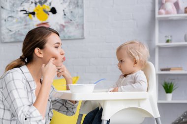 Genç bir kadın, kasenin yanındaki çocuk koltuğunda oturan çocuğa bakarken yüzüne dokunuyor.