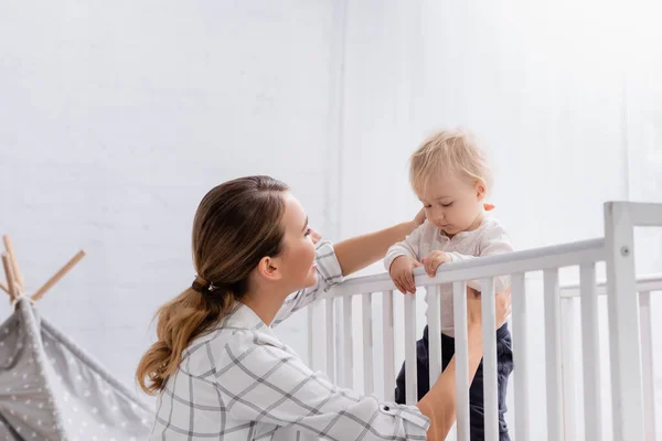 Young Mother Supporting Toddler Boy Standing Crib — Stock Photo, Image