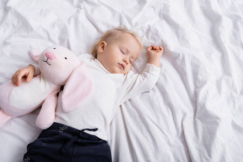 overhead view of toddler boy sleeping on white bedding with toy bunny