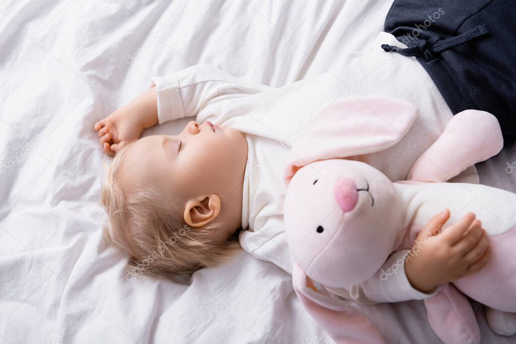 overhead view of toddler sleeping near toy bunny on white bedding