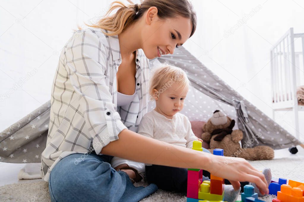 smiling woman playing with construction cubes with son on carpet near kids wigwam 