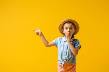 Hasır şapkalı şaşkın çocuk ağzını kapatıyor ve parmağıyla sarıyı işaret ediyor.