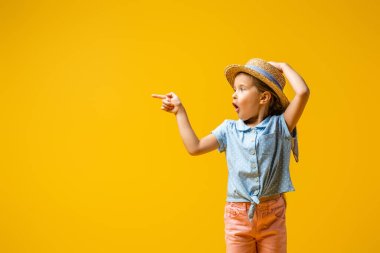 shocked kid in straw hat pointing with finger isolated on yellow clipart