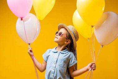 Hasır şapkalı ve güneş gözlüklü mutlu çocuk sarı balonlara bakıyor.