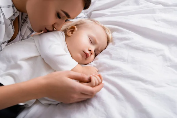 young  mother kissing sleeping son while holding his hand
