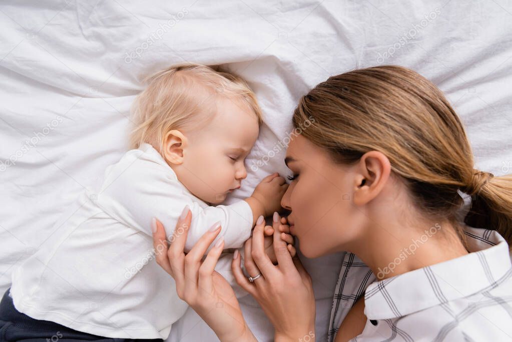 overhead view of young woman kissing hand of son sleeping on white bedding