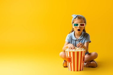 Üç boyutlu gözlüklü şaşkın çocuk patlamış mısır kovasının yanında bacak bacak üstüne atmış ve sarı üzerine film izliyor. 