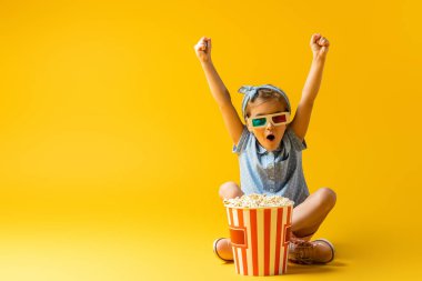 Üç boyutlu gözlüklü heyecanlı çocuk bacak bacak üstüne atmış ve patlamış mısır kovasının yanında el kaldırmış ve sarı üzerine film izliyordu.