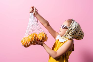 girl in headscarf and sunglasses holding reusable string bag with oranges isolated on pink clipart