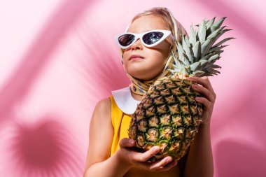 Türbanlı ve güneş gözlüklü kız pembe üzerinde ananas tutuyor. 