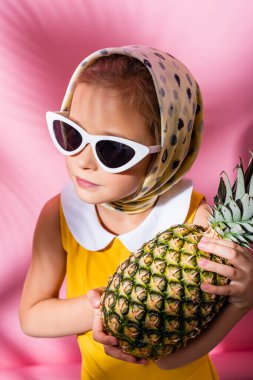 Türbanlı ve güneş gözlüklü çocuk pembe üzerinde ananas tutuyor. 