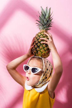 Şok olmuş başörtüsü ve güneş gözlüklü kız kafasının üstünde ananas tutuyor. 