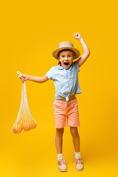 полный рост взволнованного малыша в соломенной шляпе с многоразовым шнуровым пакетом с апельсинами на желтом 