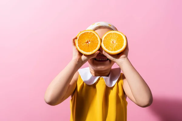 happy girl covering eyes while holding orange halves on pink