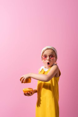 Şok olmuş başörtüsü ve güneş gözlüklü kız pembe üzerine izole edilmiş turuncu yarımlar tutuyor.