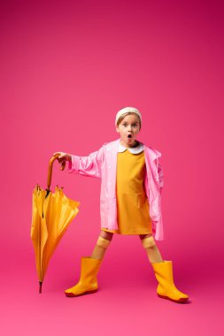 full length of shocked girl in raincoat and rain boots standing with yellow umbrella on crimson clipart