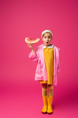 full length of shocked girl in raincoat and rain boots standing with watermelon on crimson clipart