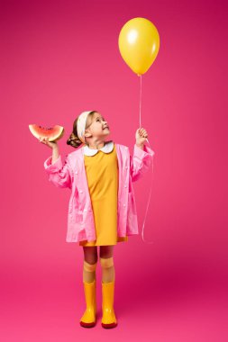 Uzun boylu, yağmurluklu, elinde balon ve kıpkırmızı kesilmiş karpuz tutan bir kız.