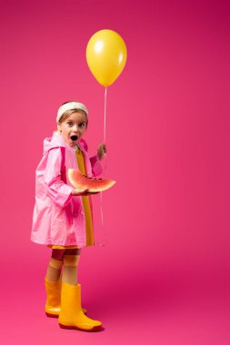 full length of amazed girl in raincoat holding balloon and sliced watermelon on crimson clipart