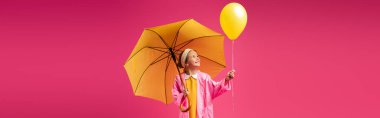 cheerful girl in raincoat holding balloon and yellow umbrella isolated on crimson, banner clipart