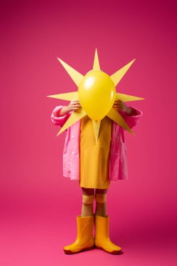 full length of girl in raincoat and rubber boots covering face while holding decorative sun with balloon on crimson clipart
